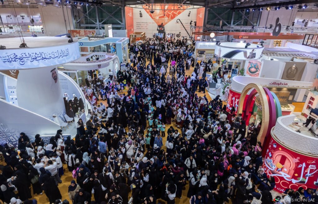 A crowded Sharjah International Book Fair 2027 with numerous stands, colorful displays, and many visitors in a large exhibition hall.