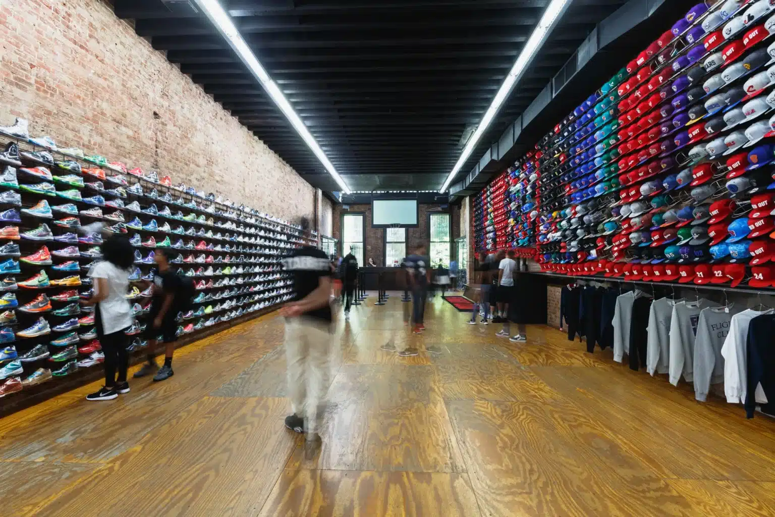 People browsing sneakers and hats on display in a store with brick walls and wooden flooring.
