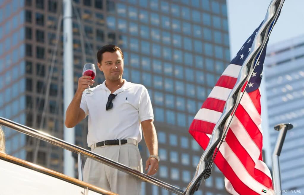 A man in a white polo shirt raises a glass on a yacht with an American flag in the foreground and city buildings behind. The Real Wolf of Wall Street