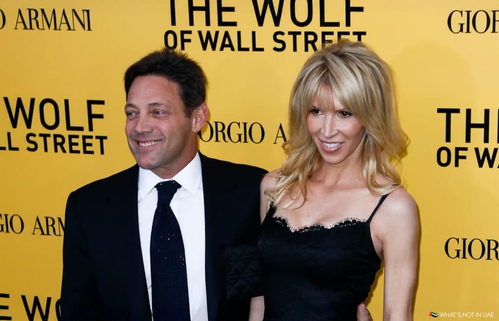 A man in a suit and a woman in a black dress pose in front of a "The Wolf of Wall Street" backdrop.