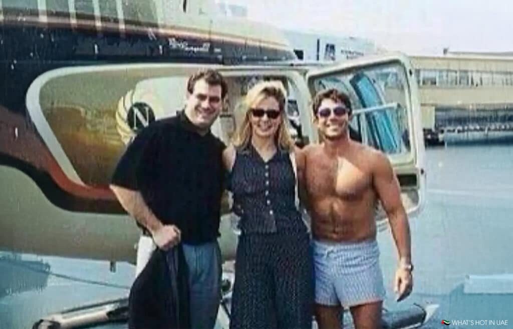 Three people smiling in front of a helicopter; one is shirtless and wearing shorts, while the others are dressed in casual attire.