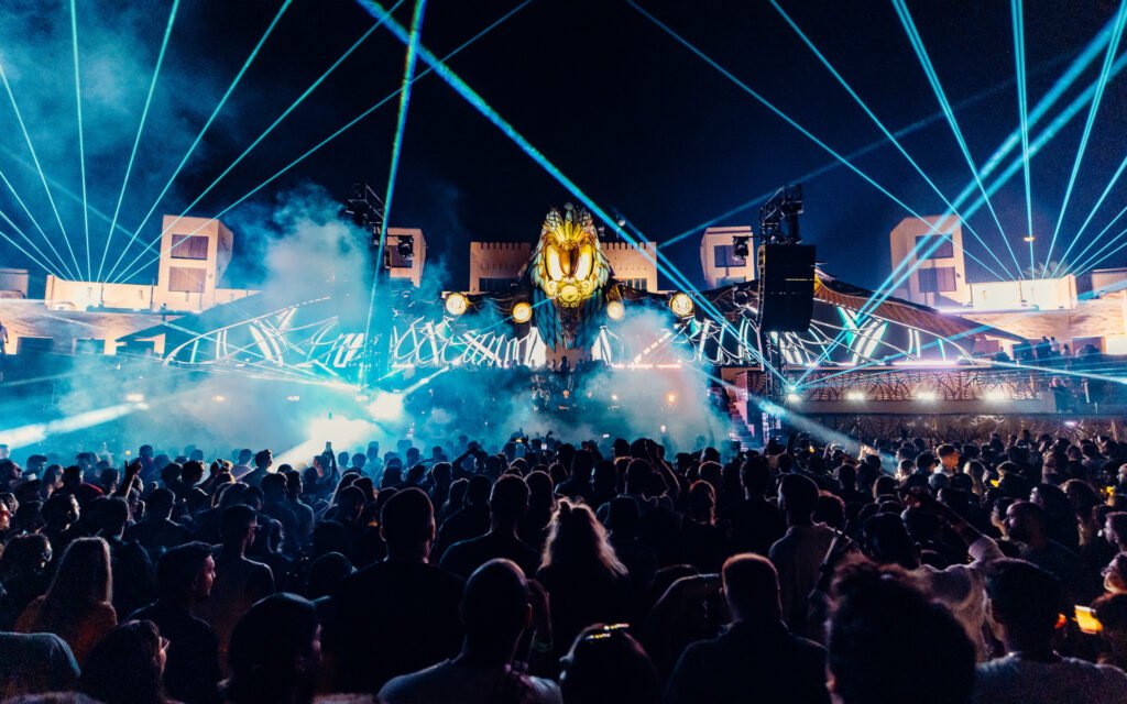 A lively festival scene with a large crowd, colorful lasers, and a stage featuring an ornate, glowing lion head.