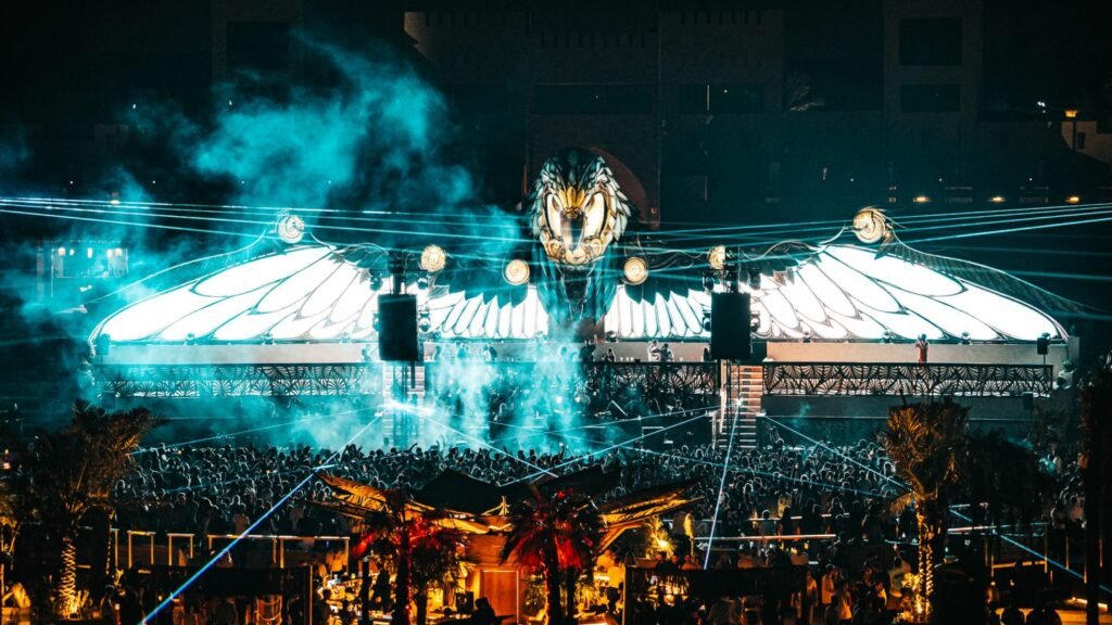 A vibrant outdoor concert at night with a large, illuminated winged stage and a crowd gathered below, surrounded by palm trees and smoke.