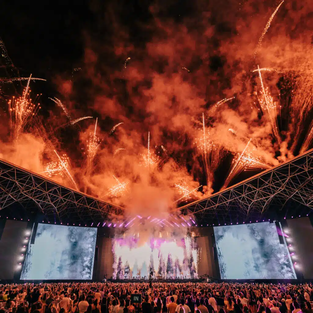 Concert stage with dramatic fireworks overhead and a large audience watching.