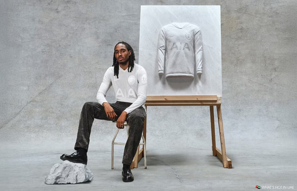 A person sits on a chair next to a marble sculpture of a jTottenham Hotspur 1901 ersey on an easel, set against a textured gray background.