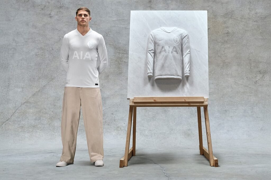 A man wearing a white Tottenham Hotspur 1901 long-sleeve shirt stands beside a stone relief sculpture of a similar shirt on an easel.
