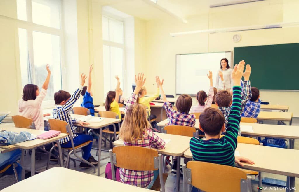 Students in a classroom raising their hands while a teacher stands at the front near a whiteboard and chalkboard.
