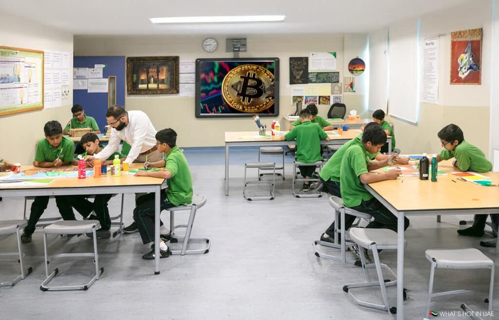 Children in green uniforms work on art projects at tables in a classroom, with a large Bitcoin image displayed on a screen.