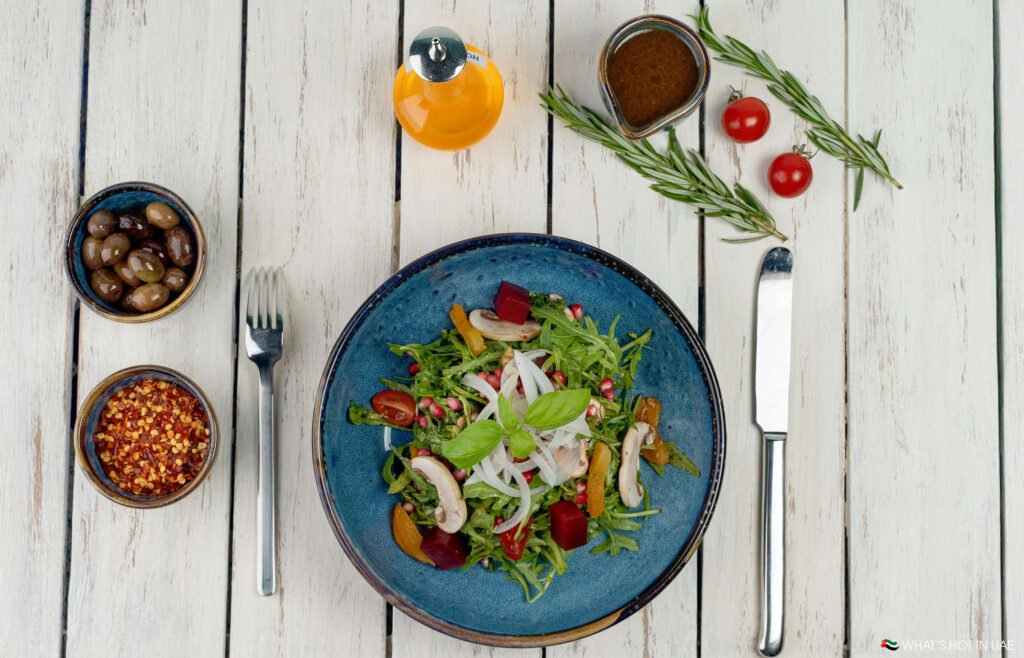 A fresh mixed salad with greens, roasted vegetables, and herbs on a blue plate, surrounded by dressing, cherry tomatoes, and seasonings on a rustic wooden table.
