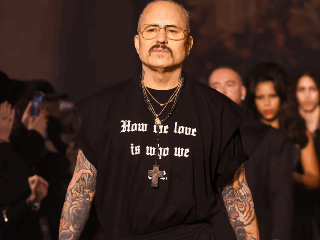 Willy Chavarria's Middle East Debut with tattoos and glasses wears a black t-shirt with text, alongside a cross necklace, on a fashion runway.