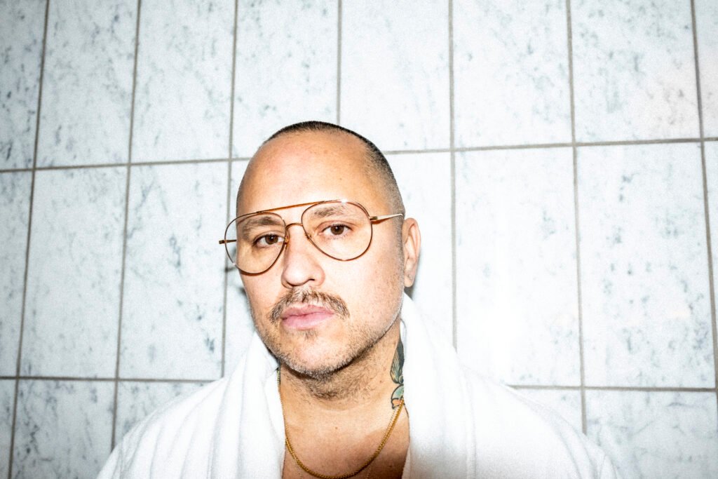 A person with glasses and short hair stands against a tiled wall, wearing a white robe.