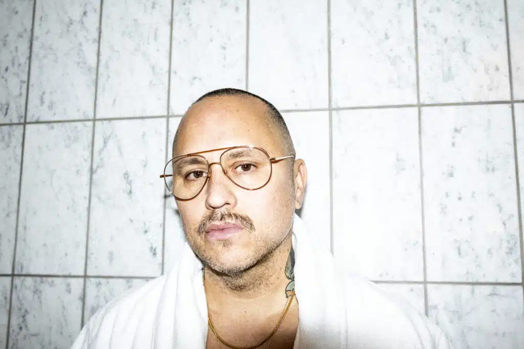 A person with glasses and short hair stands against a tiled wall, wearing a white robe.