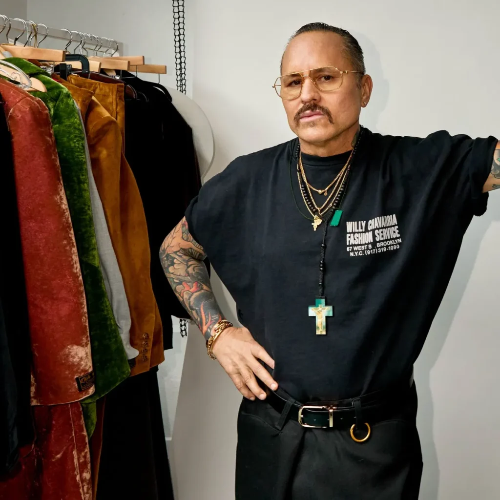 Willy Chavarria wearing a black t-shirt with "Willy Chavarria Fashion Service" is standing in a closet filled with various colorful garments. Willy Chavarria's Middle East Debut
