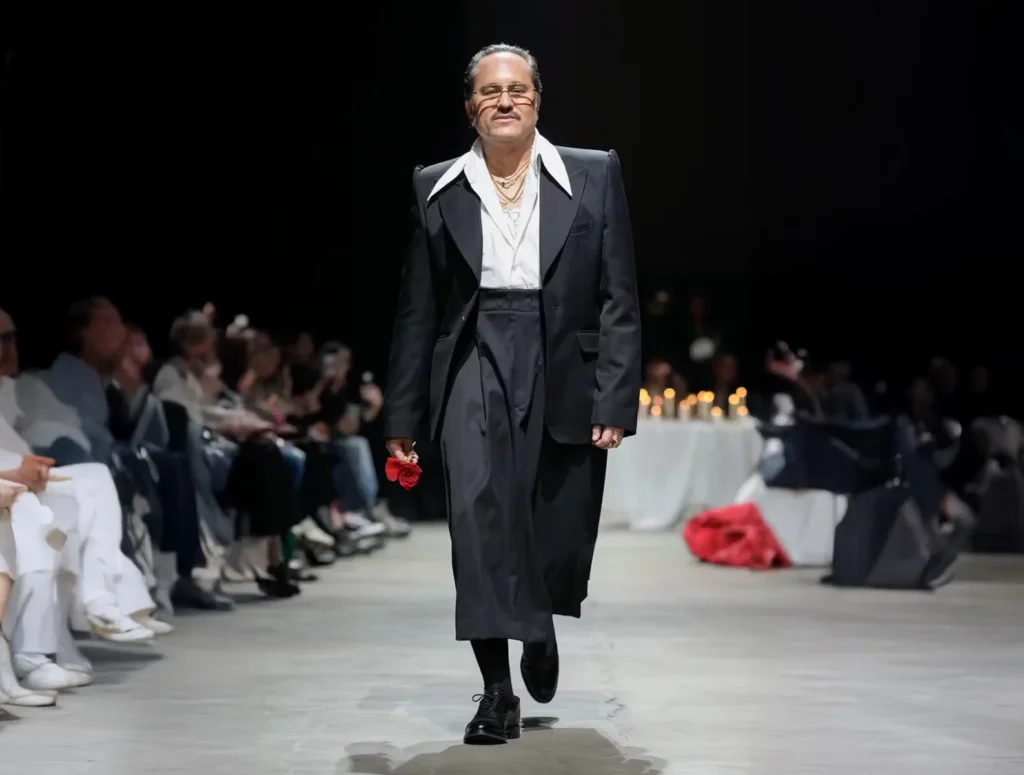 A person in formal evening attire walks confidently down a fashion runway holding a red object, with an audience seated on either side.