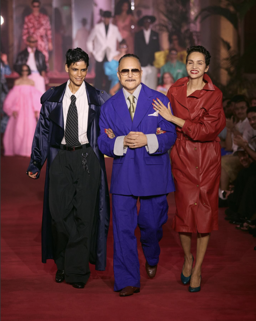 Three models on a runway, one in a blue suit and sunglasses, flanked by others in a trench coat and red leather coat.
