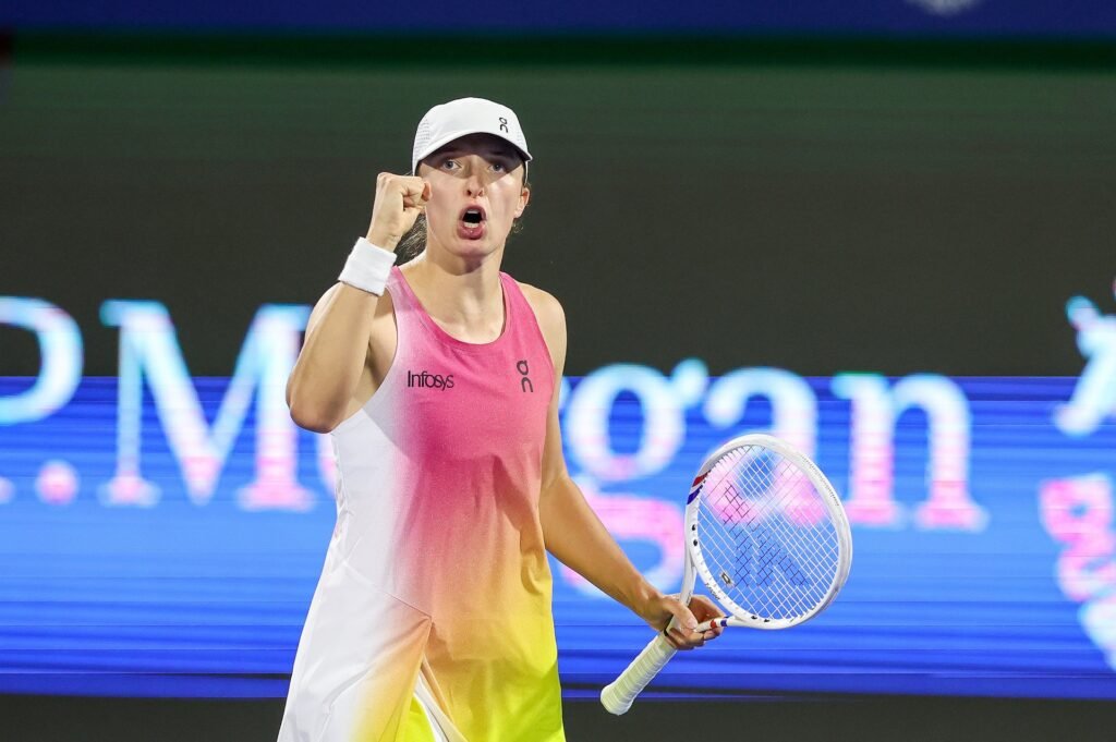 A tennis player wearing a gradient tank top and white cap raises their fist energetically, holding a racket.