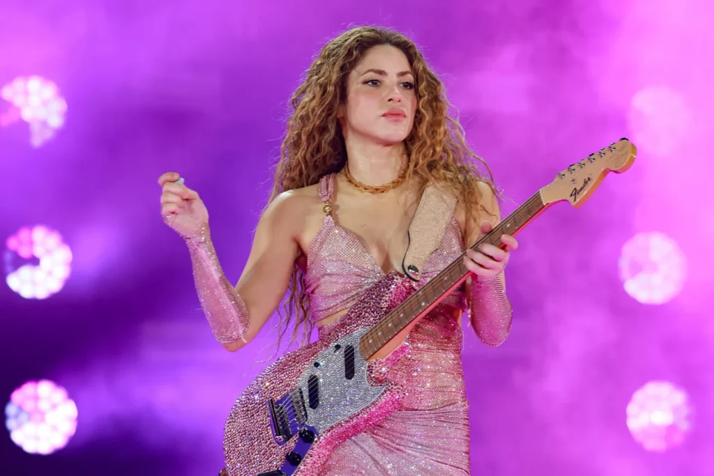 Person with curly hair in a sparkling dress playing a guitar on stage with purple lighting.