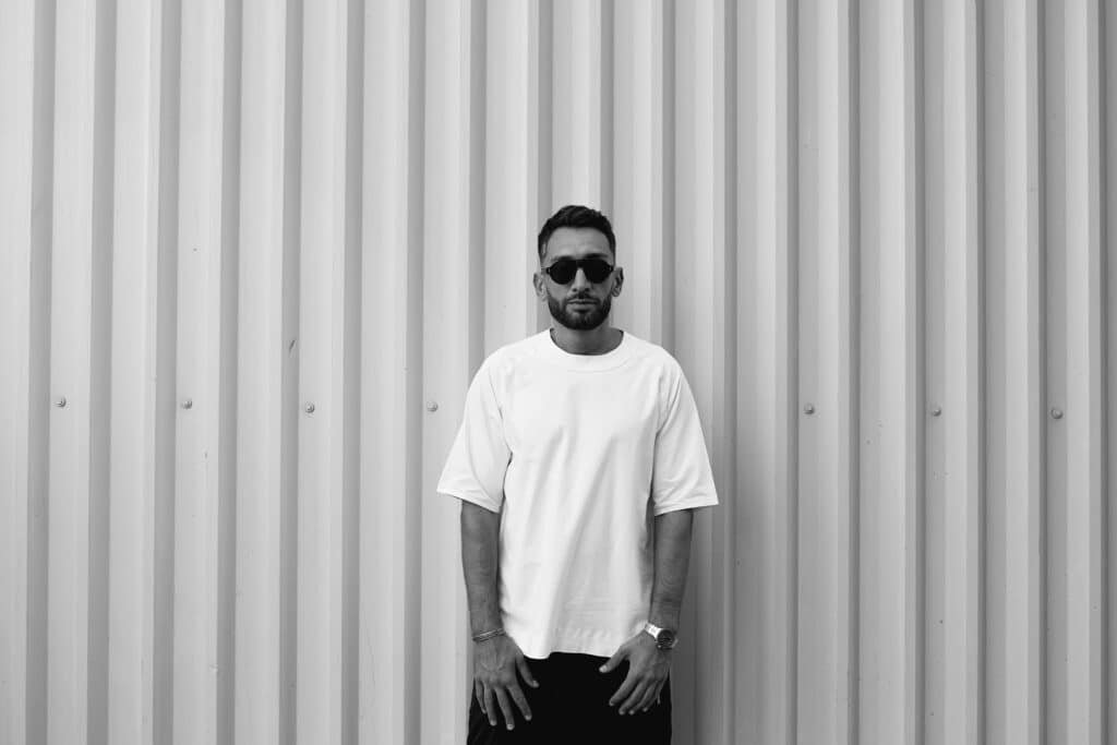 Man wearing sunglasses and a white T-shirt stands in front of a corrugated metal wall.