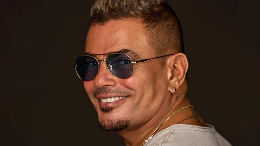 A smiling Amr Diab with sunglasses, short hair, earrings, and a goatee against a dark background.