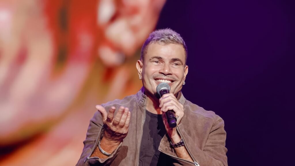 A person with short hair is smiling and holding a microphone on stage, wearing a brown jacket.