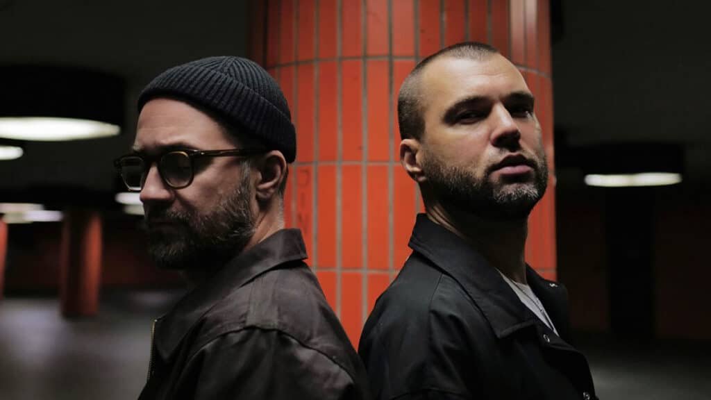 Two men stand back-to-back in a dimly lit space with orange tiled columns. One wears a beanie and glasses; the other has a shaved head.