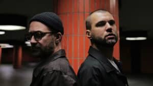 Two men stand back-to-back in a dimly lit space with orange tiled columns. One wears a beanie and glasses; the other has a shaved head.