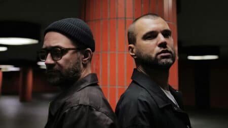 Two men stand back-to-back in a dimly lit space with orange tiled columns. One wears a beanie and glasses; the other has a shaved head.