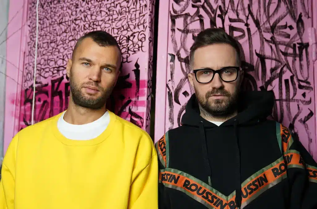 ARTBAT UPPERGROUND Ushuaïa Dubai stand side by side, one wearing a yellow sweater and the other in a black hoodie with "ROUSSIN" text, against a pink graffiti background.