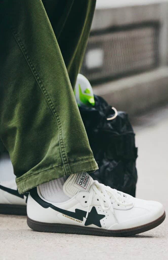 Person wearing green pants and white BAPE x adidas FIFA World Cup 26 sneakers with a black star design, sitting next to a black bag.
