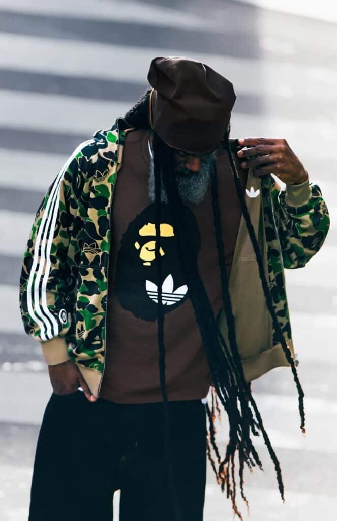 A person with long dreadlocks wearing a camouflage jacket and a T-shirt featuring a stylized BAPE x adidas FIFA World Cup 26 gorilla and Adidas logo.