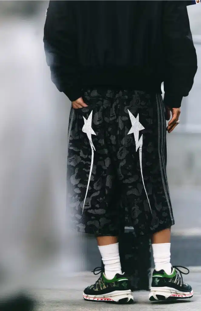 A person standing, wearing black BAPE x adidas FIFA World Cup 26 shorts with white star patterns and green sneakers with a white sole.