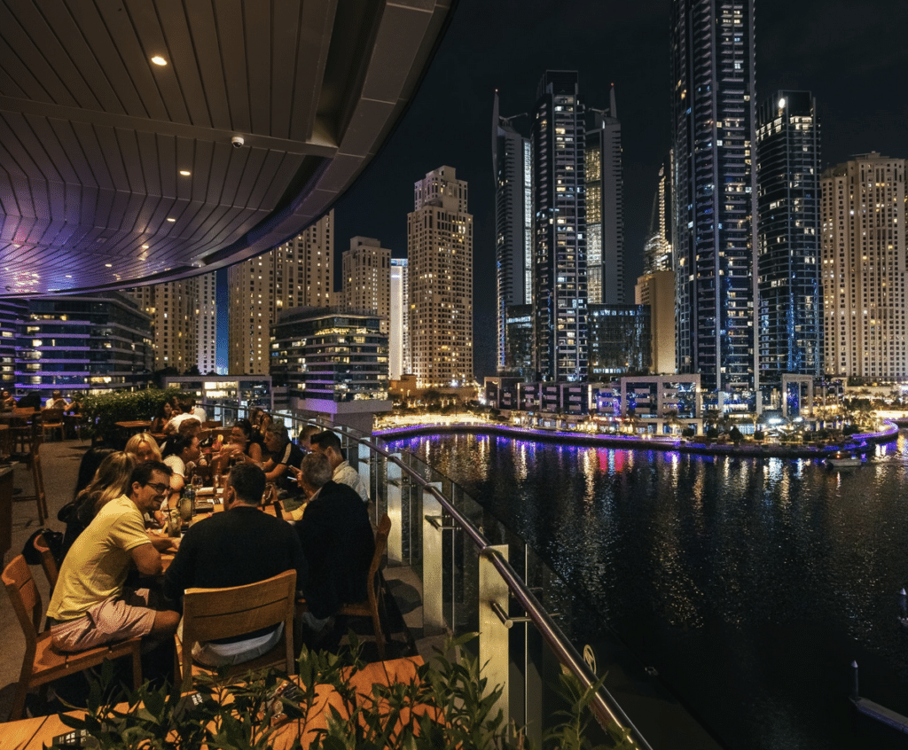 Best Bars in Dubai Marina: People dining on a balcony overlooking a city waterfront with tall, illuminated buildings at night.