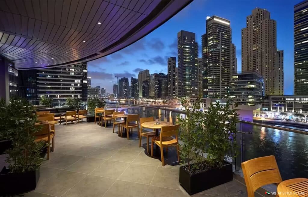 Rooftop terrace with wooden tables and chairs overlooking a city skyline and a river at dusk.