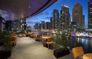 Rooftop terrace with wooden tables and chairs overlooking a city skyline and a river at dusk.