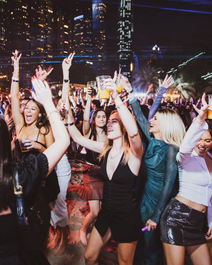 People dancing joyfully at the RONG Open Air Dubai outdoor party at night, with city skyscrapers lit up in the background.