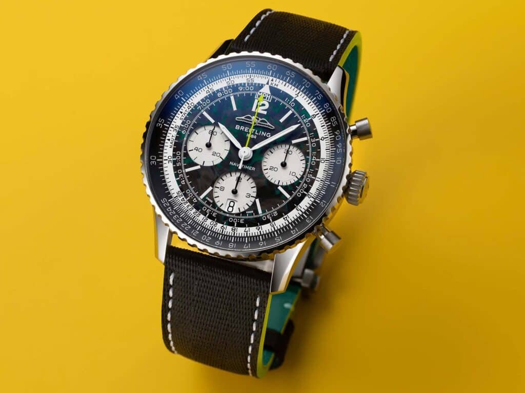 A luxury Breitling Navitimer Aston Martin watch with a black strap, detailed dial, and silver casing against a vibrant yellow background.