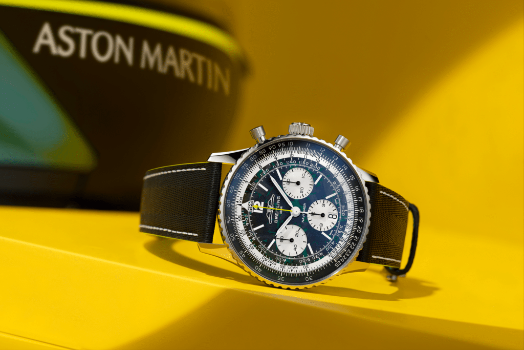A luxury wristwatch with a black strap and chronograph features, set against a yellow background with partially visible Aston Martin text.