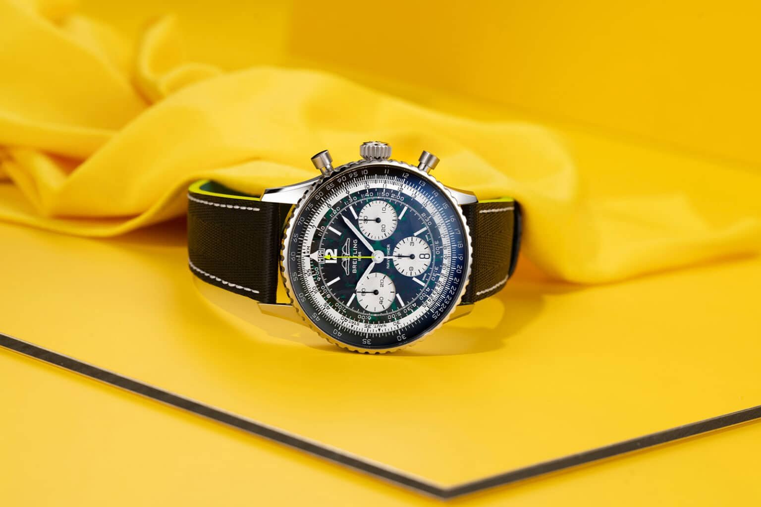 A silver and black chronograph watch with a leather strap is displayed against a bright yellow background.