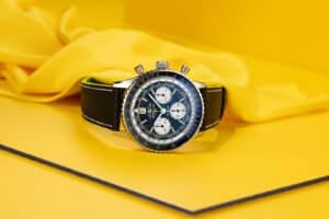 A silver and black chronograph watch with a leather strap is displayed against a bright yellow background.