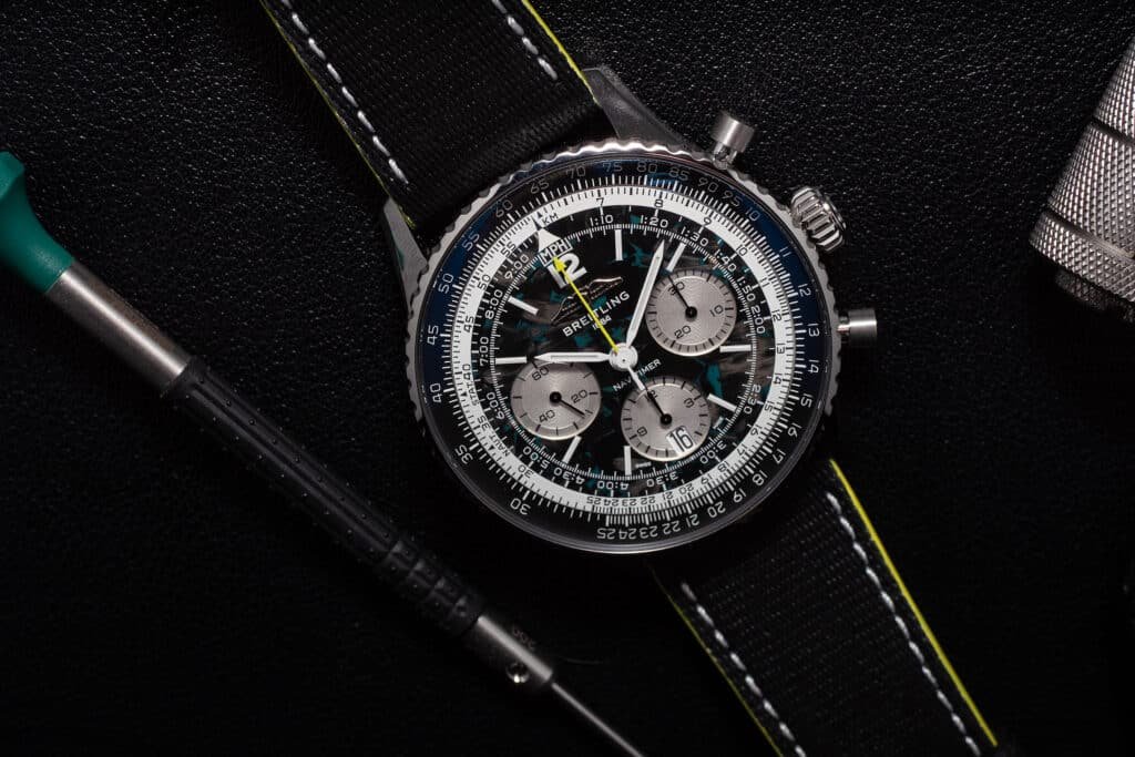 A Breitling Navitimer Aston Martin chronograph watch with a dark strap and intricate dial, placed on a textured black surface.