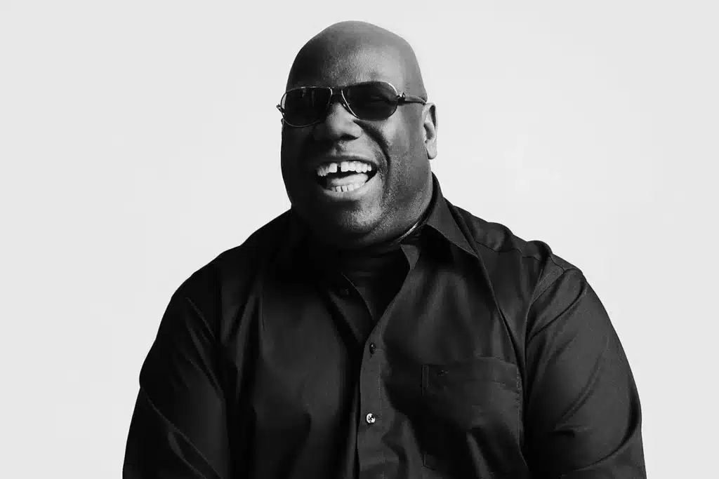 Carl Cox wearing sunglasses and a dark shirt is smiling broadly in a black and white photo.