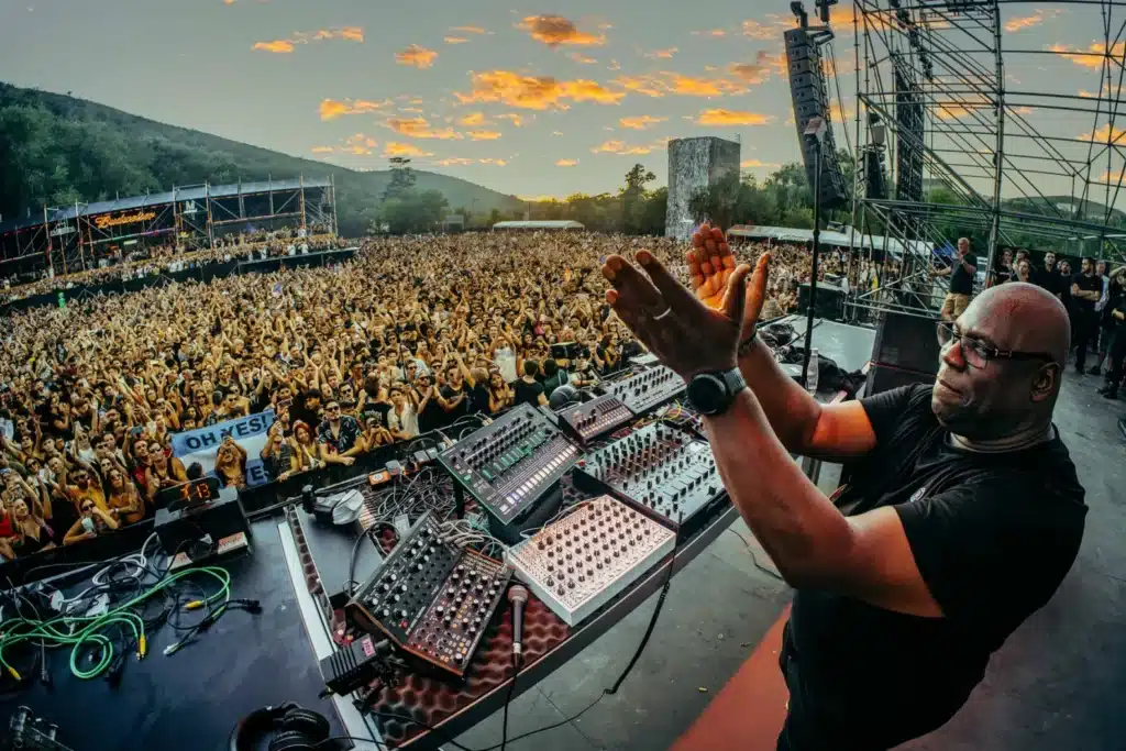 DJ Carl Cox performing on stage, clapping with a cheering crowd and scenic sunset in the background.