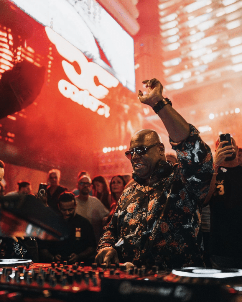 Booking a Global DJ in Dubai like Carl Cox with sunglasses and a floral shirt performs energetically on stage in front of a crowd, with colorful lights in the background.
