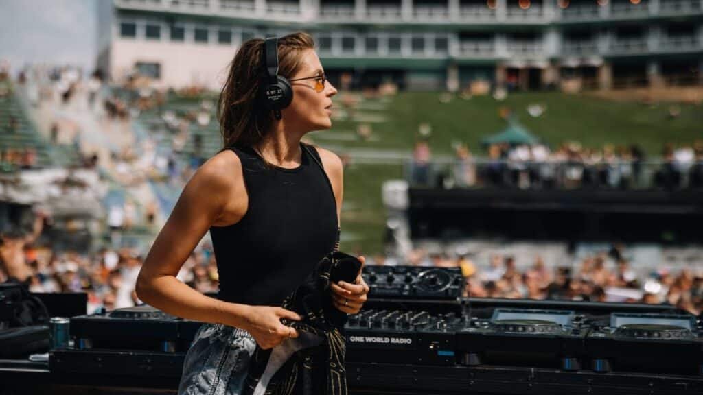 Charlotte de Witte wearing headphones stands at a mixing table outdoors, with a large crowd and grassy area in the background.
