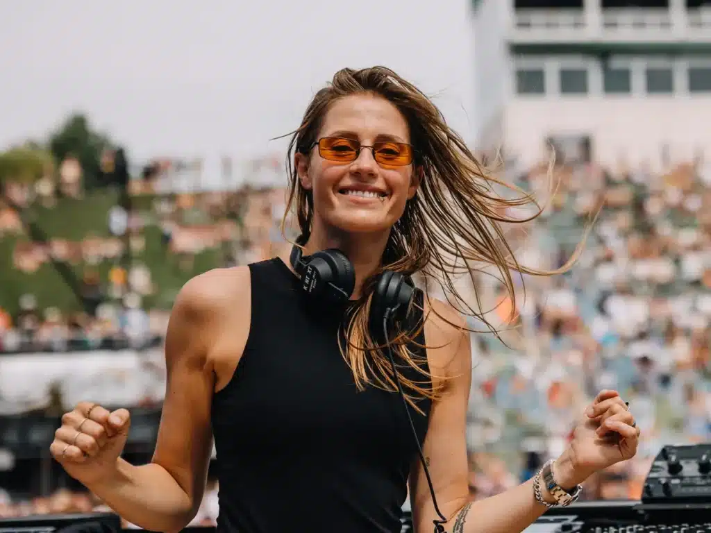 A smiling Charlotte de Witte with long hair wearing amber sunglasses and headphones, standing outdoors at an event.