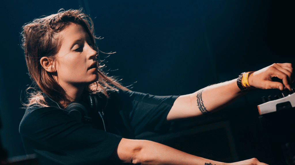 Charlotte de Witte at HIVE with headphones is focused on adjusting audio equipment on stage under dim lighting.
