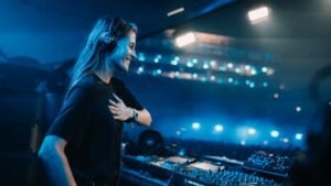 A DJ wearing headphones is performing on stage at a concert with blue lighting and a large audience in the background.