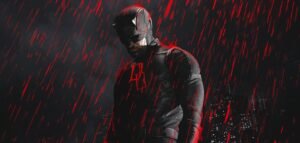 A superhero in a dark suit with horns and a red emblem stands under red rain against a cityscape.