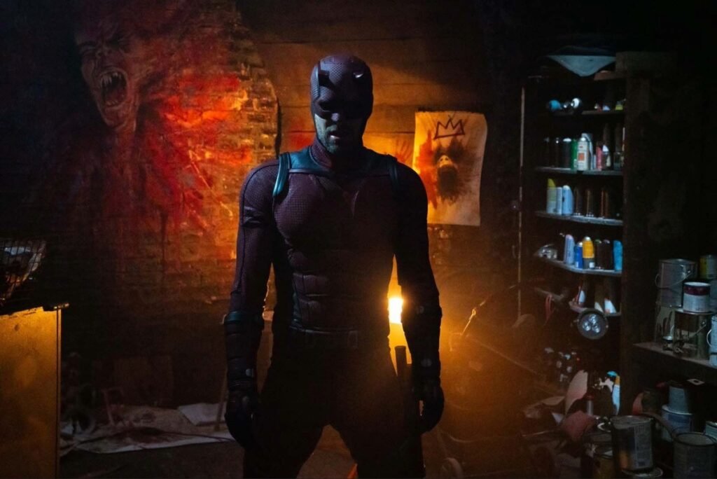 A person in a dark, textured superhero suit stands in a dimly lit room with paintings and shelves of paint supplies in the background.