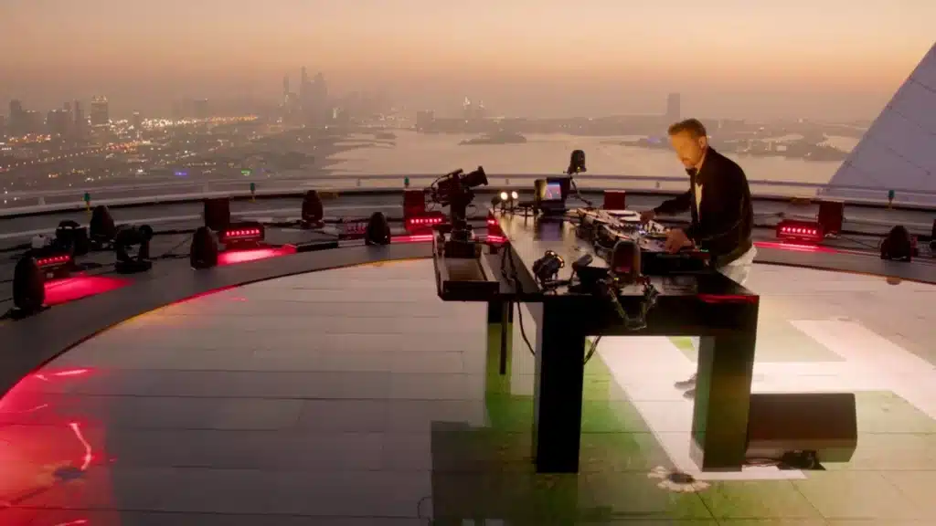A DJ performing on a rooftop at sunset with a city skyline and water in the background.